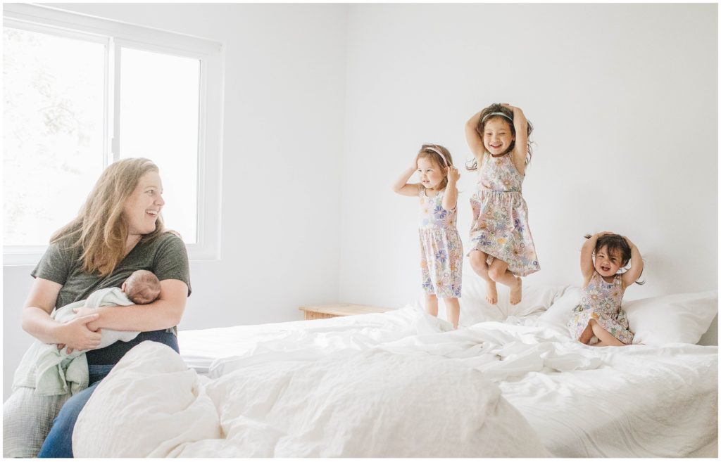Image of siblings jumping on the bed while mom holds newborn baby brother